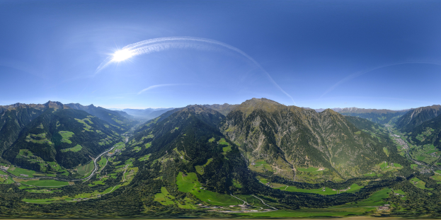 Panoramischer Blick auf ein Gebirgsmassiv von einem Hügel aus, mit grünem Gras, einer gewundenen Straße und einem hellblauen Himmel mit Sonnenlicht.