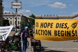Eine Gruppe von Menschen steht vor einem Gebäude und hält ein gelbes Banner mit der Aufschrift "Hope Dies, Action Begins", während in der Nähe Fahrräder abgestellt sind und ein Pfahl mit einer Tafel daran steht, vor einem klaren blauen Himmel mit Bäumen im Hintergrund.