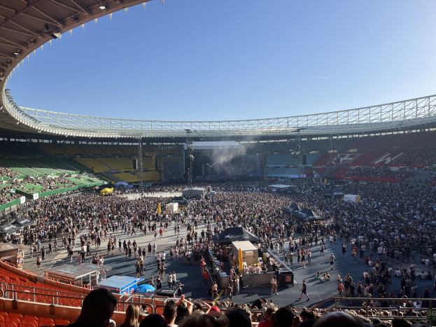 Eine große Menschenmenge füllt ein Stadion mit einer zentralen Bühne, sichtbarem Himmel und Dachbeleuchtung, was auf ein Konzert im Wembley-Stadion hinweist.
