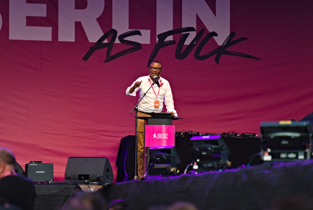 Ein Mann in einem weißen Hemd und braunen Hosen steht auf einer Bühne und hält ein Mikrofon, mit einem pinken Banner im Hintergrund, auf dem 'Berlin as Fuck' steht, und Lautsprechern auf der Bühne.