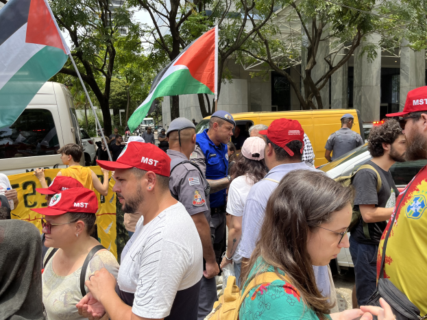Eine Gruppe von Menschen steht vor einem Gebäude, einige tragen Mützen und andere Taschen, halten palästinensische Flaggen, mit Fahrzeugen auf der Straße und Bäumen im Hintergrund unter einem klaren blauen Himmel.