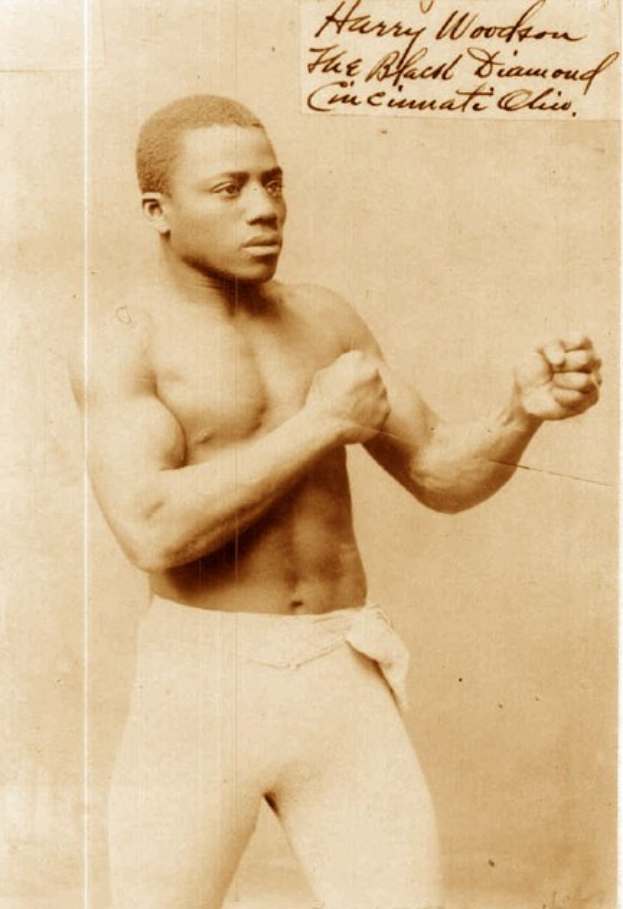 Ein Schwarz-Weiß-Foto eines Mannes in einem Boxring mit Handschuhen und Shorts, der mit entschlossener Miene und konzentriertem Blick dasteht.