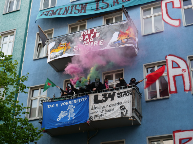 Eine Gruppe von Menschen auf einem Balkon mit Flaggen und Transparenten, mit einem Baum links und Rauch in der Luft, was auf eine Demonstration hindeutet.