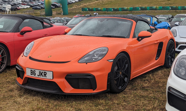 Porsche 718 Boxster GTS auf dem Goodwood Festival of Speed, umgeben von anderen Autos auf einer grünen Fläche mit einem Banner, Bäumen und einem klaren blauen Himmel.
