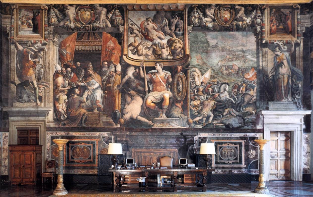 Ein großer Raum mit einem prominenten Gemälde an der Wand, ein Tisch mit Gegenständen, zwei Lampen, zwei Türen und ein Teppich, befindet sich im Palazzo della Signoria in Florenz, Italien.