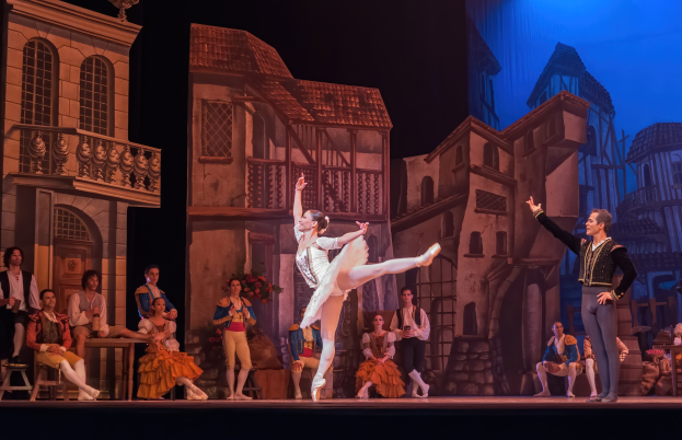 Zwei Tänzer performen auf einer Bühne im Royal Opera House in London, mit einem Publikum im Vordergrund, das auf Stühlen sitzt und steht, und einem Bildschirm im Hintergrund, der Gebäude anzeigt.