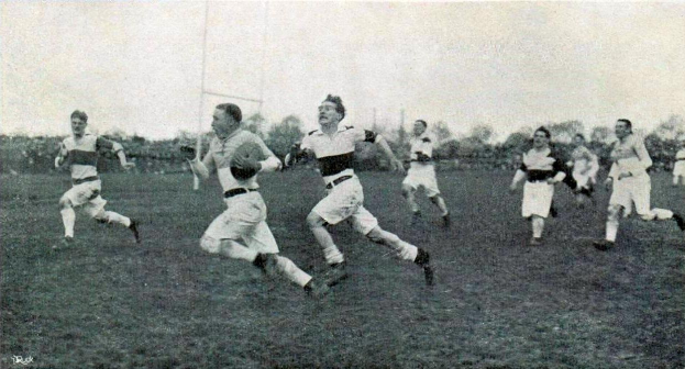 Schwarz-weiß-Foto von Männern, die Rugby auf einem Rasenfeld in den 1930er Jahren spielen, mit Bäumen und Pfählen im Hintergrund unter einem klaren Himmel.