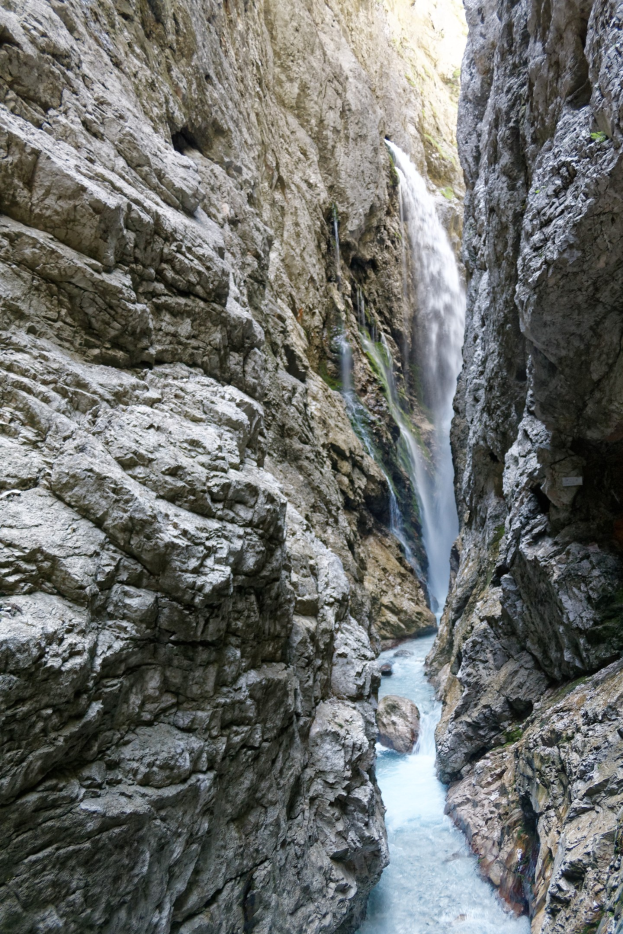 Ein kleiner Wasserfall ergießt sich über zerklüftete Felsen in einem steinigen Tal, umgeben von saftig grünen Hügeln, mit kristallklarem Wasser, das von hellerer Sonne beleuchtet wird.
