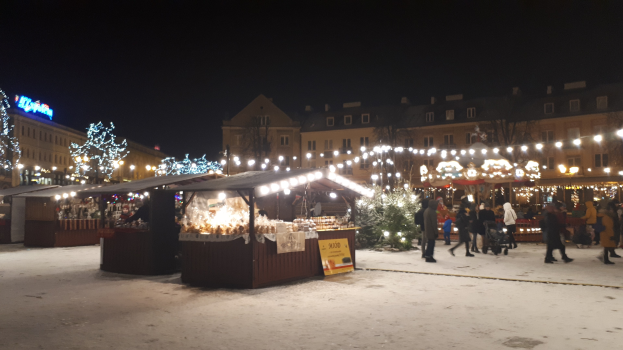 Lebendiger Weihnachtsmarkt an einem verschneiten Abend mit Menschen, Buden, Pflanzen, Bäumen, Gebäuden, Schildern und einem bewölkten Himmel.