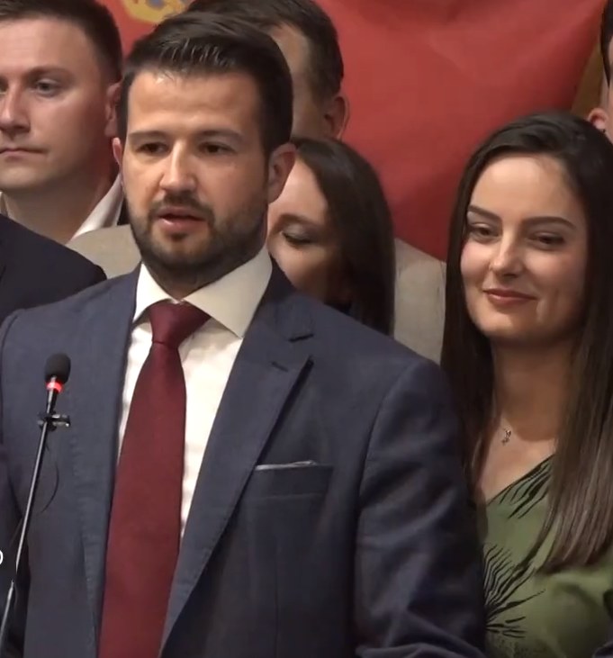 Ein Mann im Anzug spricht vor einem Publikum an einem Rednerpult, mit einem Banner dahinter und einer Menge davor.