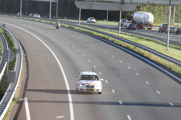 Ein Auto auf einer Autobahn mit einem stationären Polizeiwagen am Rande, andere Fahrzeuge, Leitplanken, Pfosten, Schilder und Bäume im Hintergrund.
