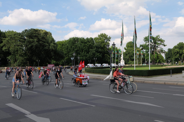 Eine Gruppe von Menschen, die auf Fahrrädern eine von Bäumen gesäumte Straße mit Laternenmasten und Fahnenmasten entlangfahren, während der Pariser Radtour 2016, mit Fahrzeugen und einer bewölkten Kulisse im Hintergrund.