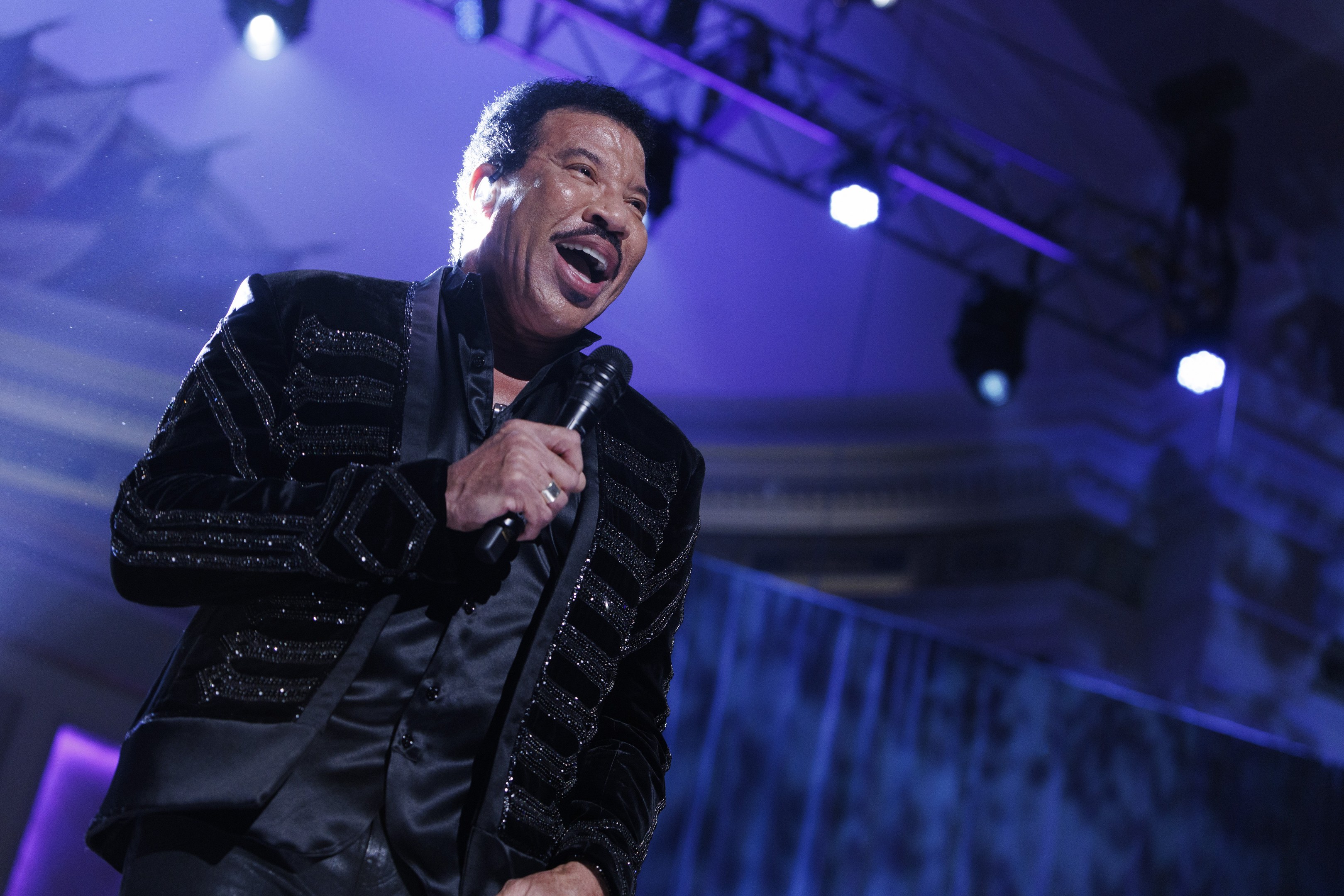 Lionel Richie performs bei den Grammys, trägt ein schwarzes Kleid und hält ein Mikrofon auf einer hell erleuchteten Bühne.