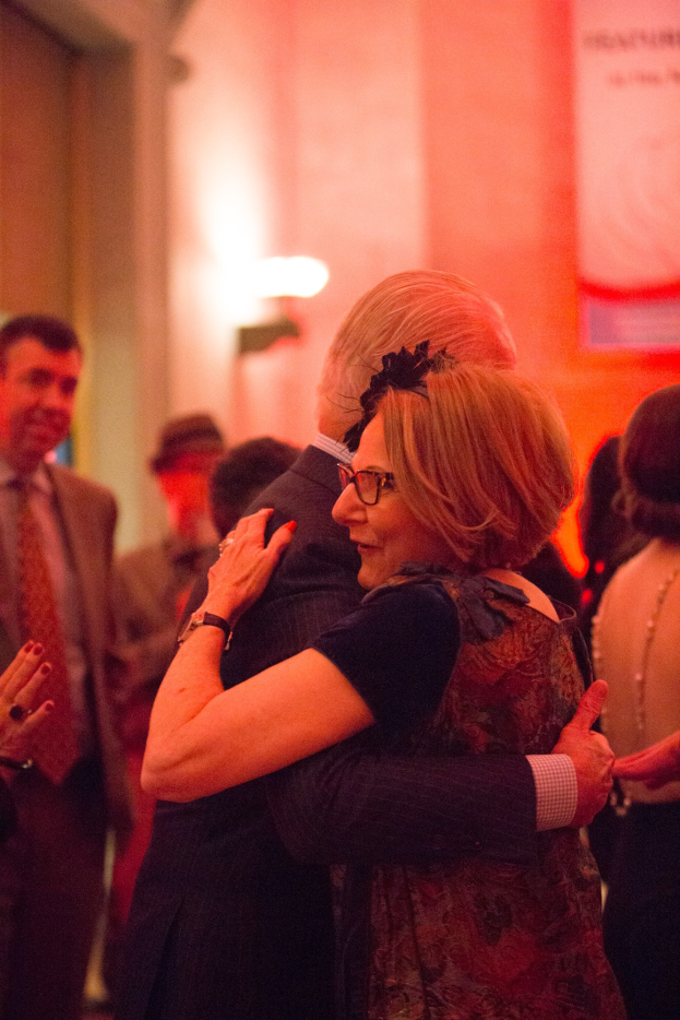 Ein Mann und eine Frau umarmen sich herzlich auf einer Party, umgeben von anderen Menschen und einer beleuchteten Wand im Hintergrund.