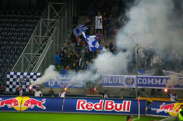 Gruppe von Menschen auf einem Fußballfeld mit Fahnen und Schildern, die Rauch aus dem Boden aufsteigen lassen und von einem Metallzaun umgeben sind, in einem Stadion mit leeren Stühlen auf der linken Seite.