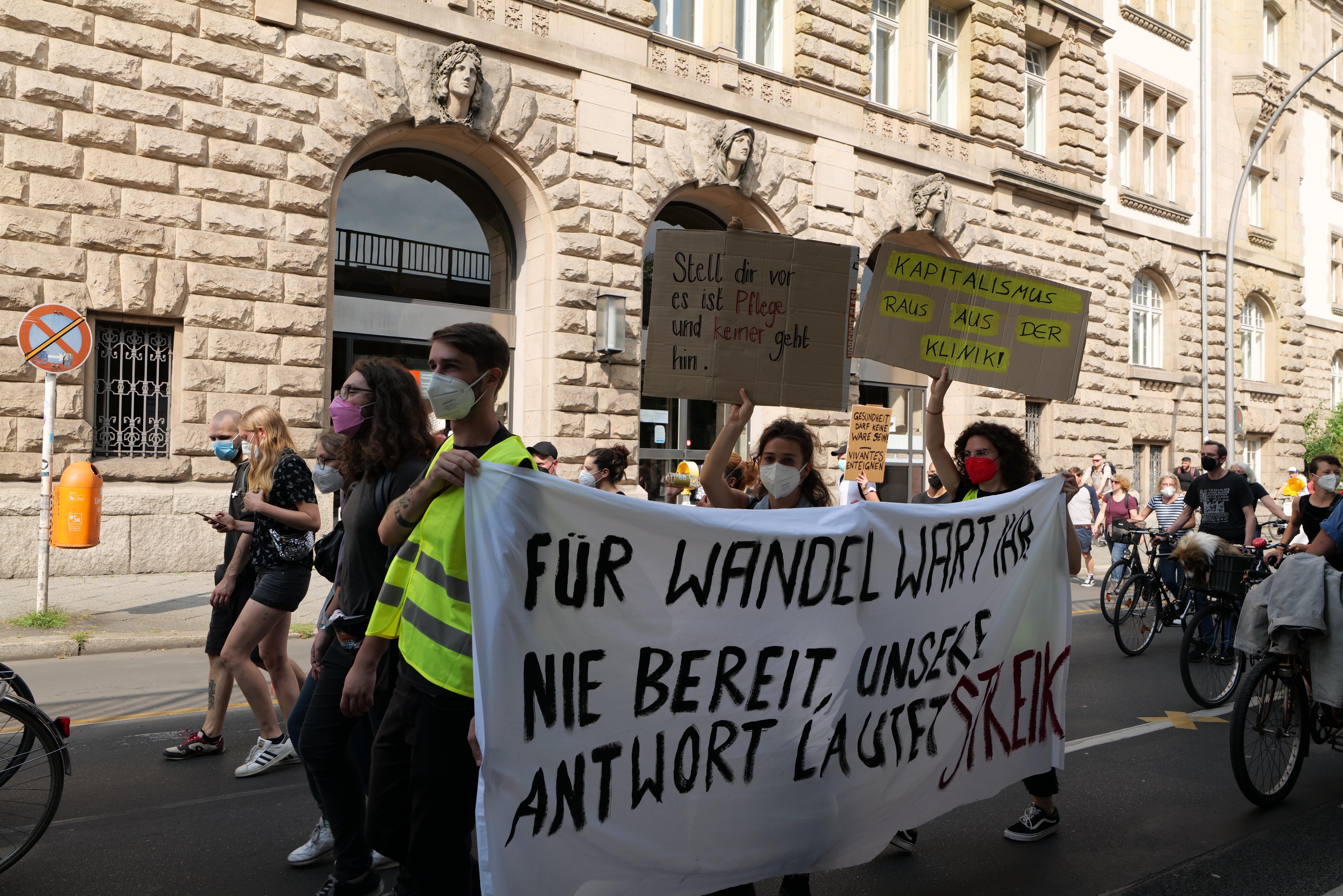 Eine Gruppe von Menschen marschiert bei einer Demonstration auf einer Stadtstraße, einige halten Schilder und andere fahren Fahrräder, mit einem historischen Gebäude mit Rundbogenfenstern, Säulen und Skulpturen im Hintergrund.