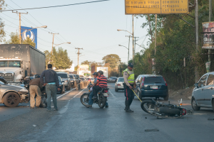 Eine Gruppe von Menschen steht um ein verunglücktes Motorrad auf der Straße mit mehreren Fahrzeugen, darunter ein Lastwagen, und einer Hintergrund von Bäumen, Pfählen, Lampen und Schildern unter einem Himmel.