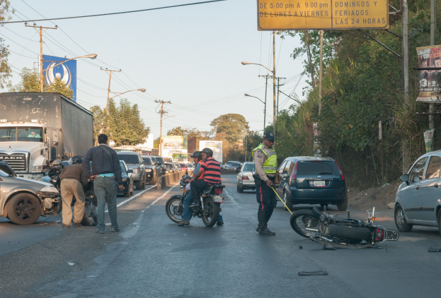 Eine Gruppe von Menschen steht um ein verunglücktes Motorrad auf der Straße mit mehreren Fahrzeugen, darunter ein Lastwagen, und einer Hintergrund von Bäumen, Pfählen, Lampen und Schildern unter einem Himmel.