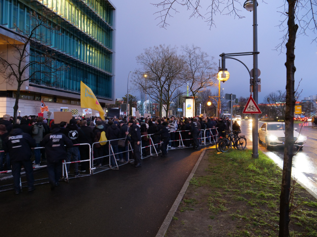 Eine große Gruppe von Menschen protestiert vor einem Gebäude in Berlin, hält Schilder, mit Barrikaden, Fahrrädern, Laternenmasten, Schildern, Bäumen und Gras im Hintergrund.