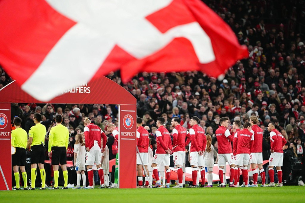 Gruppe von Menschen auf einem Fußballfeld mit einer roten und weißen Flagge im Vordergrund, einem Bogen mit 'Bayern Munich vs Bayern Munich Betting Tips & Preview' im Hintergrund und einer großen Menschenmenge im Stadion.