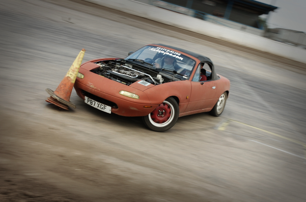 Eine Person fährt ein rotes Auto auf einer Rennstrecke vorbei an einem Verkehrskegel mit einem unscharfen Hintergrund, der hohe Geschwindigkeit andeutet.