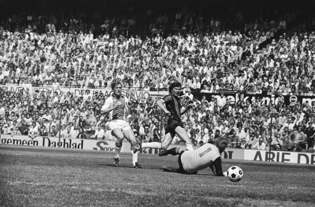 Schwarzes Foto von Männern, die auf einem Feld Fußball spielen, mit Zuschauern im Hintergrund und einer 'Bundesliga 1978-79 Ajax 1-1 Hannover 96'-Fahne.