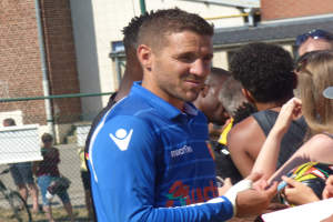 Ein Mann in einem blauen Hemd signiert Autogramme für eine Gruppe von Menschen, einige tragen Kappen und Schutzbrillen, mit einem Gebäude, Maschendrahtzaun und einem Fahrrad im Hintergrund.