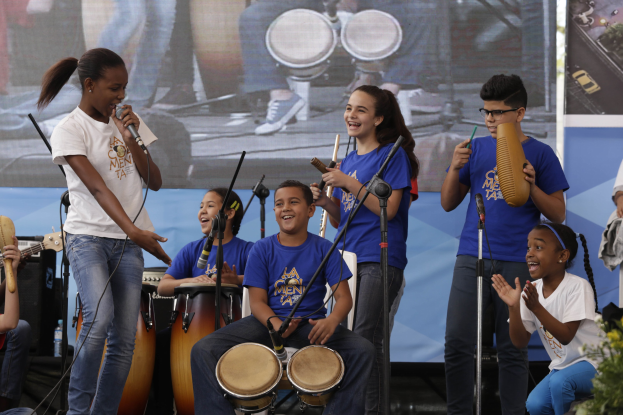 Eine Gruppe von Kindern, die auf einer Bühne Musikinstrumente spielen, mit einer Frau, die ein Mikrofon hält, umgeben von Mikrofonstativen, einem Banner im Hintergrund und Pflanzen auf der rechten Seite.