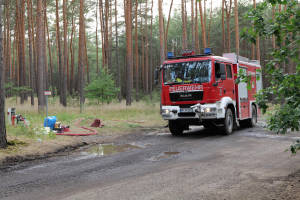 Ein rotes Feuerwehrauto fährt eine Schotterstraße entlang, umgeben von Bäumen, Gras, einem Rohr und anderen Gegenständen auf der linken Seite der Straße, sowie weiteren Bäumen und einem klaren blauen Himmel im Hintergrund.