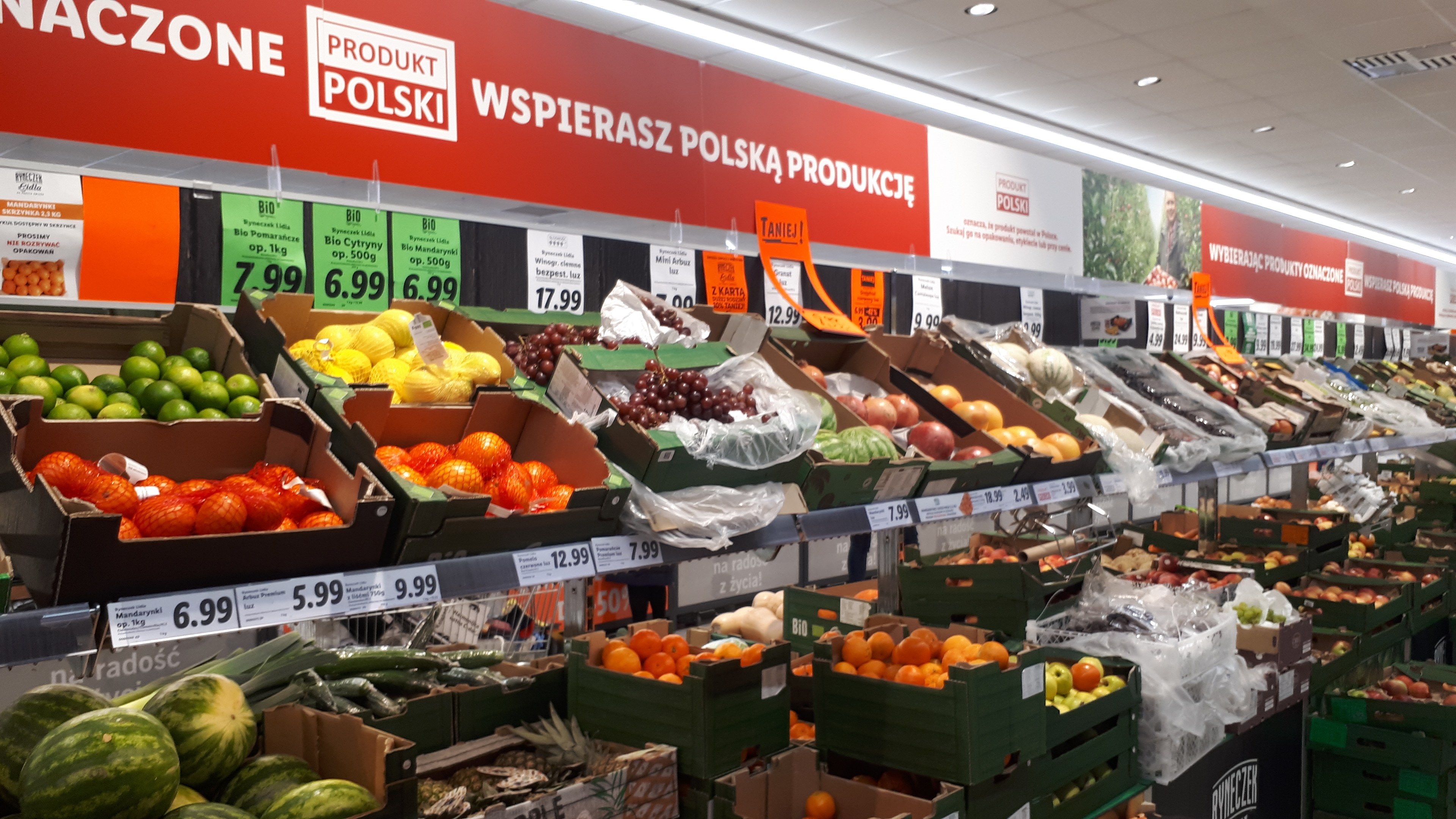 Ein Geschäftsraum mit verschiedenen Obst- und Gemüsesorten in Kartons mit Preisschildern, Wandtafeln mit Text und Bildern sowie Deckenbeleuchtung.