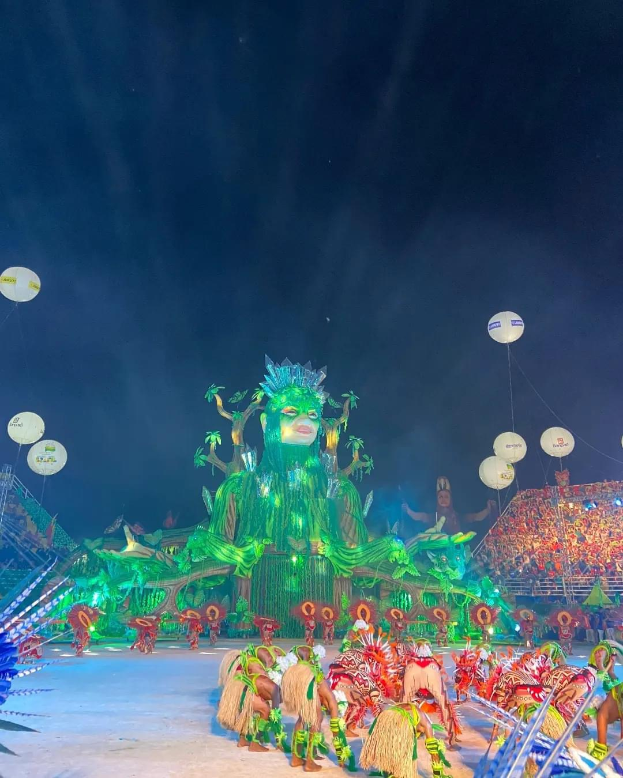Ein farbenfroher Karneval in Rio de Janeiro, Brasilien, mit Performern in bunten Kostümen auf der Bühne, einer großen Menge im Publikum und einem Himmel voller farbiger Lichter und Ballons.