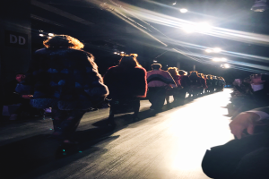 Eine Gruppe von Menschen läuft auf einem Laufsteg bei einer Fashion Show, beleuchtet von hellen Scheinwerfern, während Zuschauer auf beiden Seiten zusehen.