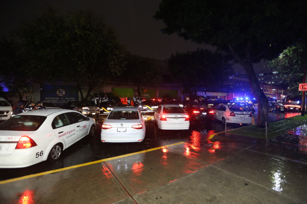 Eine Gruppe von Polizeiwagen auf einem Parkplatz bei Nacht mit sichtbarer Beschilderung, Gras, Bäumen, Gebäuden, einem Banner, Lichtern und einer bewölkten Himmel.