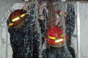 Drei Feuerwehrleute in schwarzen Uniformen und roten Helmen stehen in einem wassergeschädigten Raum und arbeiten zusammen, um ein Feuer zu löschen.
