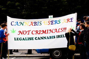 Eine Gruppe von Menschen auf der Straße mit einem Banner, auf dem "Cannabisnutzer sind keine Kriminellen" steht, mit einem parkenden Fahrzeug in der Nähe und Bäumen und einem Zaun im Hintergrund.