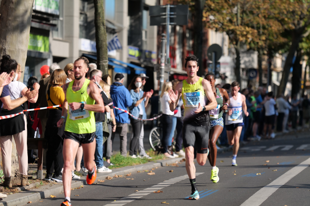 Gruppe von Läufern bei einem Marathon auf einer Stadtstraße, mit Zuschauern auf dem linken Gehweg, Bäumen, Gebäuden und einem klaren blauen Himmel im Hintergrund.
