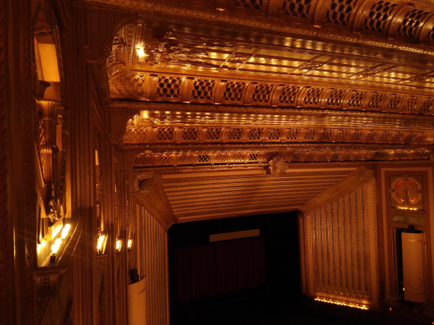 Ein prächtiger Theaterinnenraum mit dunklem Hintergrund, beleuchtet von links und rechts, mit einer kunstvoll gestalteten Decke.