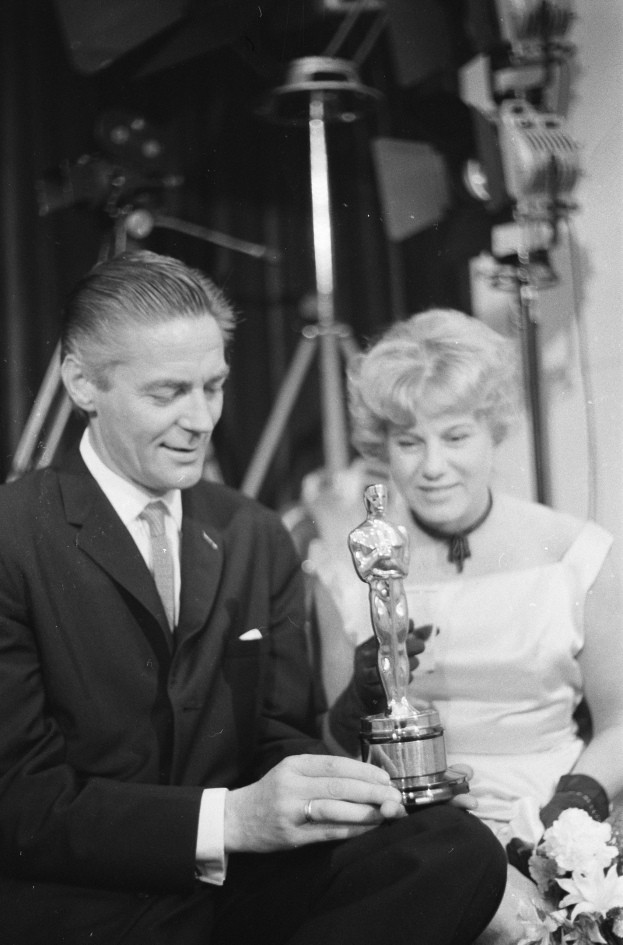 Ein Schwarz-Wei├č-Bild eines formell gekleideten Mannes, der eine Oscar-Statue und eine Frau mit einem Blumenstrau├čus in den Händen hält, sitzend nebeneinander mit beleuchteter Hintergrund.