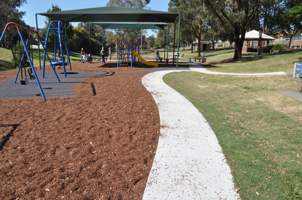 Ein Park mit einem Spielplatz, der Wippen, Rutschen und andere Spielgeräte bietet, umgeben von grünem Gras, Bäumen, Bänken, Schuppen im Hintergrund und einem klaren blauen Himmel.