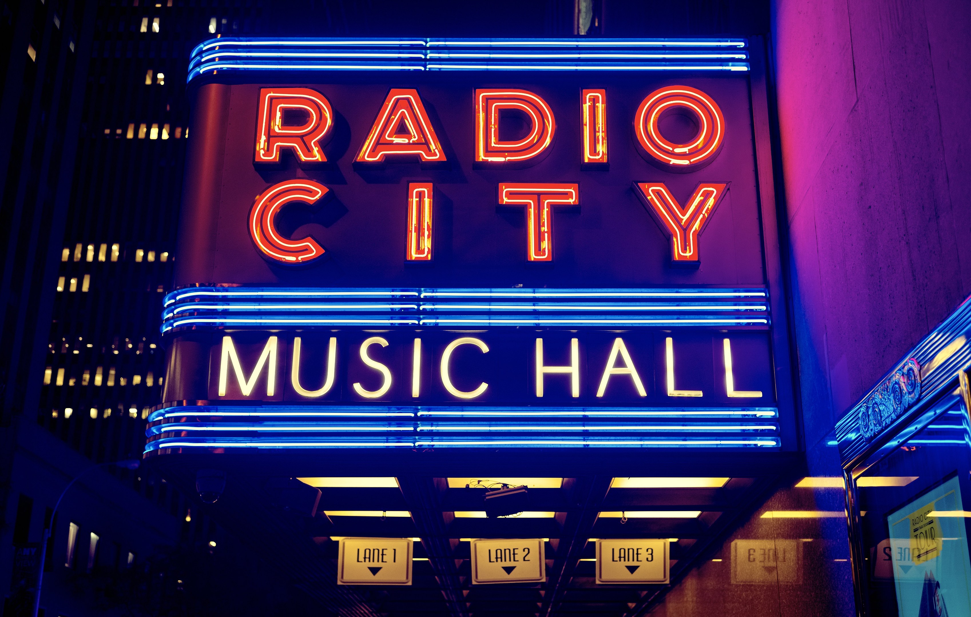 Außenansicht des Radio City Music Hall in New York City bei Nacht, hell erleuchtet mit hohen Gebäuden im Hintergrund und einem "Radio City"-Schild auf der rechten Seite.