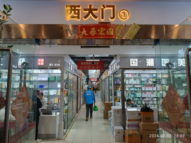Innenraum einer chinesischen Apotheke mit Glaswänden, Regalen voller Artikel, verstreuten Kartons und einer Pflanze auf der linken Seite; ein Schild oben weist darauf hin, dass es sich um eine Drogerie handelt.