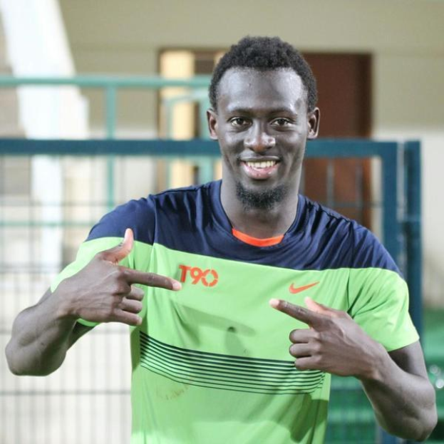 Ein Mann in einem grünen und blauen Hemd, der in die Kamera lächelt und auf sie zeigt, mit einem unscharfen Hintergrund aus einem Geländer, einer Wand und einer Tür.