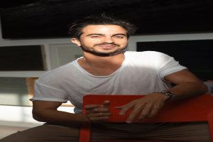 Ein Mann sitzt auf einem roten Stuhl in einem Raum, lächelt, trägt ein weißes T-Shirt und eine Uhr, mit einer Wand und einem Fenster im Hintergrund.