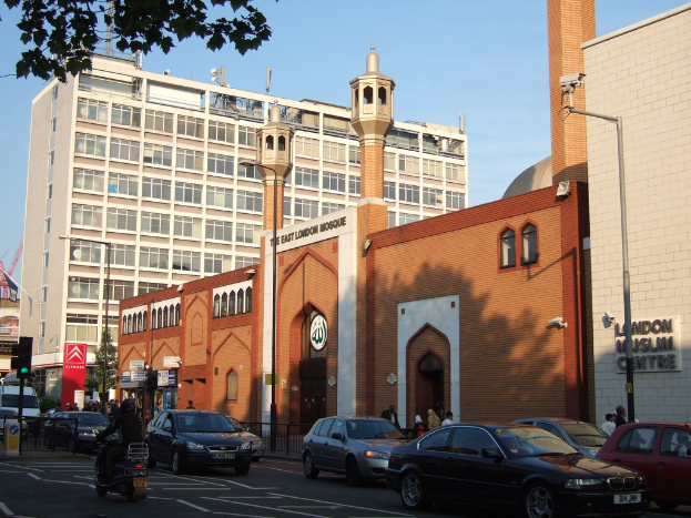 Eine Moschee mit einem Namenbrett an der Wand, Straßenlaternen, eine Ampel an einem Mast, Fahrzeuge auf der Straße, Gruppen von Menschen, die stehen, ein Baum und eine Überwachungskamera an einem Mast.
