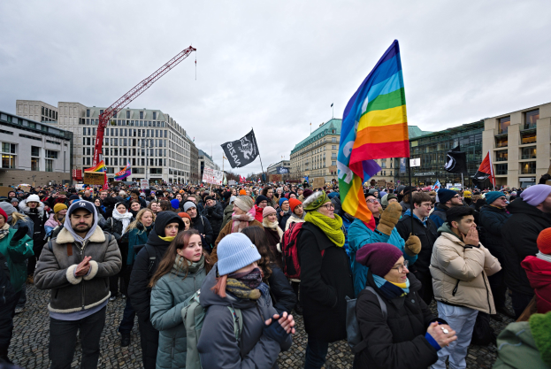 Eine große Gruppe von Menschen mit Fahnen und Schildern mit Text, darunter "Lgbtq+ Rechte Demonstration in Berlin", vor einem Gebäude mit einem Kran und einem bewölkten Himmel im Hintergrund.