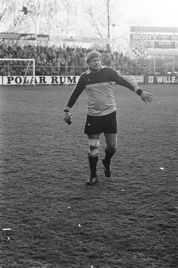 Ein professioneller Fütballer in einem Fütballtrikot steht auf einem Feld umgeben von Zuschauern, einem Tor, Brettern und Bäumen unter einem klaren Himmel.