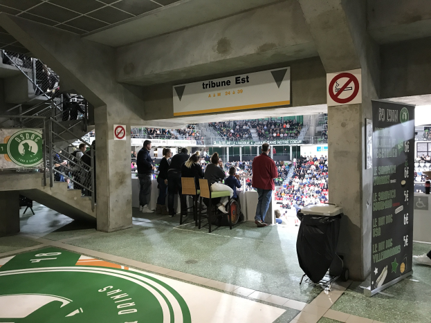 Eine Gruppe von Menschen steht vor einem sitzenden Publikum in einem Stadion, mit Schildern, die "Boston Celtics vs New York Jets", ein Müllcontainer, eine Tafel mit Text, eine Treppe mit Geländern und ein Dach mit Deckenleuchten zeigen.