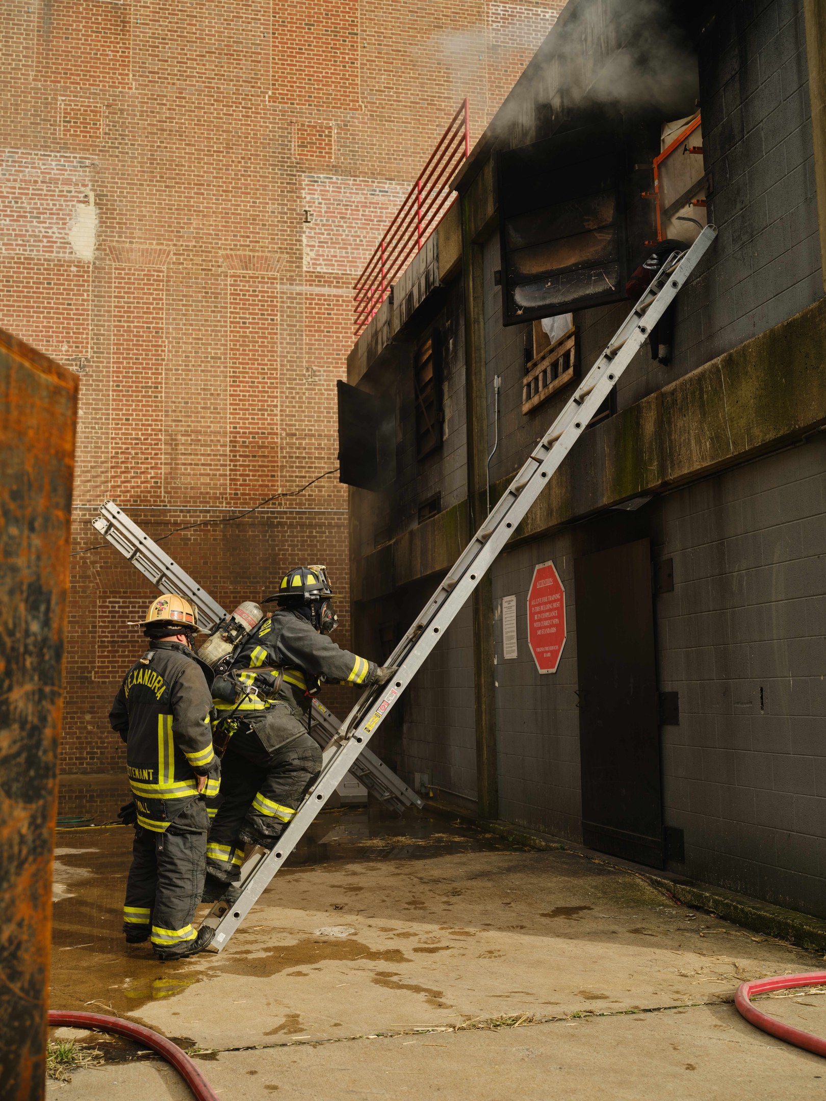 Feuerwehrleute in Helmen, die eine Leiter zu einem rauchgefüllten Gebäude hinaufsteigen, mit einem Metallobjekt links und Rohren auf dem Boden.