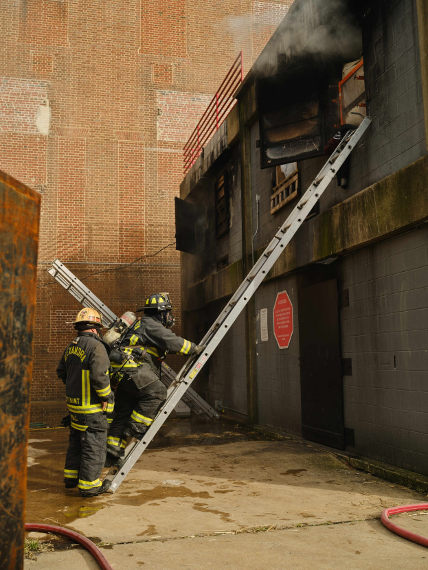 Feuerwehrleute in Helmen, die eine Leiter zu einem rauchgefüllten Gebäude hinaufsteigen, mit einem Metallobjekt links und Rohren auf dem Boden.