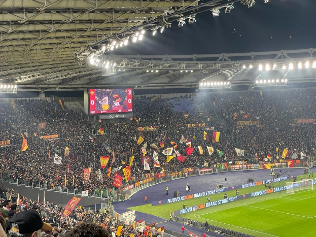 Eine große Menge Menschen in einem Stadion schwenkt Fahnen, mit Gras, einem Netz, Bannern und einem Bildschirm zu sehen; Menschen in Mützen füllen die Tribüne unter beleuchteten Lichtern, was Spannung vor einem Fußballspiel vermuten lässt.